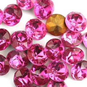 May include: A collection of pink, faceted, round, plastic rhinestones. The rhinestones are scattered on a white surface.