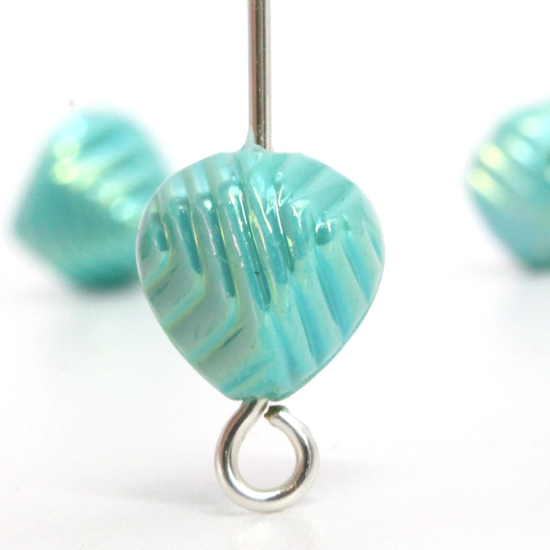 May include: Close-up of a turquoise leaf-shaped bead with a textured surface and a silver-colored eye pin. The bead has a pearlescent sheen and is in focus, with two other beads slightly blurred in the background. The beads are likely for jewelry making.