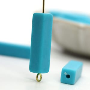 May include: Two turquoise blue rectangular polymer clay beads with a gold jump ring on one bead. The beads are smooth and have a matte finish.