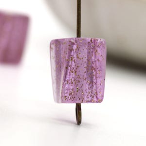 Puede incluir: Una sola cuenta de vidrio cuadrada de color morado claro con purpurina dorada incrustada en el vidrio. La cuenta está en un alambre.