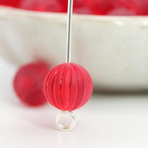 May include: A close-up of a red, round bead with vertical grooves, strung on a silver-colored wire. The bead has a loop at the bottom. Other red beads are visible in the background, near a white bowl.