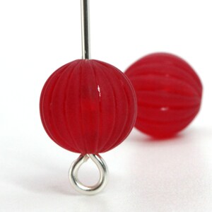 May include: Close-up of two red, round beads with a ribbed texture. One bead is attached to a silver-colored metal pin with a loop, while the other is slightly out of focus in the background. The beads have a glossy finish.