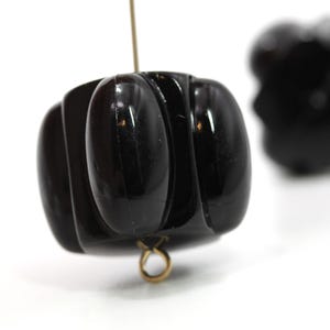 May include: Close-up of a pair of black, carved, vintage earrings. The earrings have a ribbed, rounded design and are suspended from gold-colored loops. The background is white, highlighting the dark, glossy finish of the earrings.