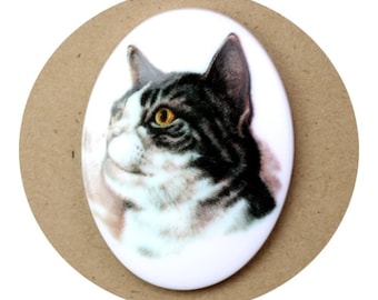 May include: A white oval ceramic tile with a black and white cat with yellow eyes looking to the right.