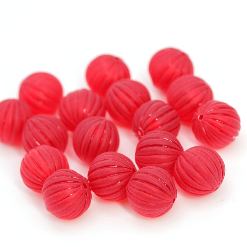 May include: A collection of red, round beads with a grooved surface. The beads are scattered on a white background. These beads could be used for jewelry making or other craft projects.