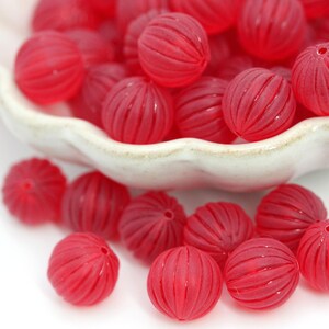 May include: A collection of translucent red, round beads with grooved surfaces. Some beads are spilling out of a white, scalloped-edge bowl. The beads have a hole for stringing and are scattered on a white surface.