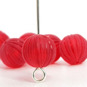 May include: Close-up of several red, ribbed glass beads with a silver-colored eye pin. The beads are spherical and have a translucent quality. The beads are likely for jewelry making or other craft projects.