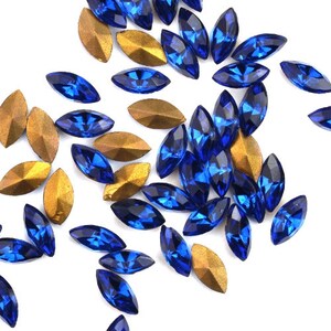 Op de afbeelding: Assortiment van blauwe glazen strasssteentjes in een marquise-vorm. De strasssteentjes zijn verspreid over een witte achtergrond.