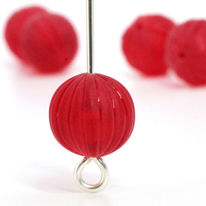 May include: Close-up of a red, round bead with vertical grooves, attached to a silver-colored eye pin. The bead is in focus, with other blurred red beads in the background. The beads are likely for jewelry making.