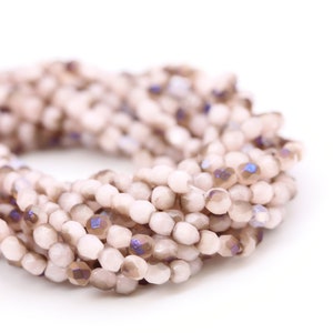 May include: A circular strand of small, faceted, opaque light pink glass beads. The beads feature a smooth, rounded shape with multiple flat facets that catch the light, interspersed with occasional iridescent purple and brown accents.
