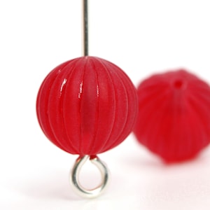 May include: Close-up of a red, ribbed bead with a silver-colored loop attached. Another red bead is slightly out of focus in the background. The beads have a glossy finish and are likely for jewelry making.