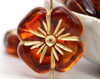 May include: Close-up of a flower-shaped bead with a golden center and accents. The bead is a translucent amber color, with a golden wire loop for stringing. The flower design has a starburst pattern in the center.