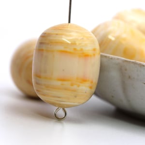 May include: Close-up of a cream-colored, barrel-shaped bead with orange and yellow striations. The bead is strung on a wire with a silver-colored loop at the bottom. Other beads are visible in the background, along with a ceramic bowl.