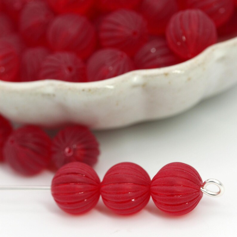May include: Close-up of red, ribbed glass beads. Three beads are strung on a wire, with a silver loop at the end. A white bowl filled with similar beads is in the background. The beads have a round shape and a textured surface.