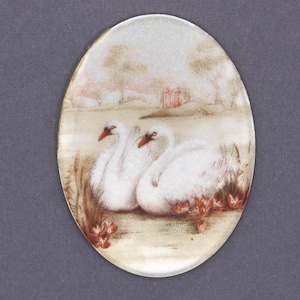 May include: An oval-shaped, white porcelain cameo with a painted scene of two white swans in a pond with brown reeds and pink flowers. The background is a light brown with a faint pink sky and a small brown building in the distance.