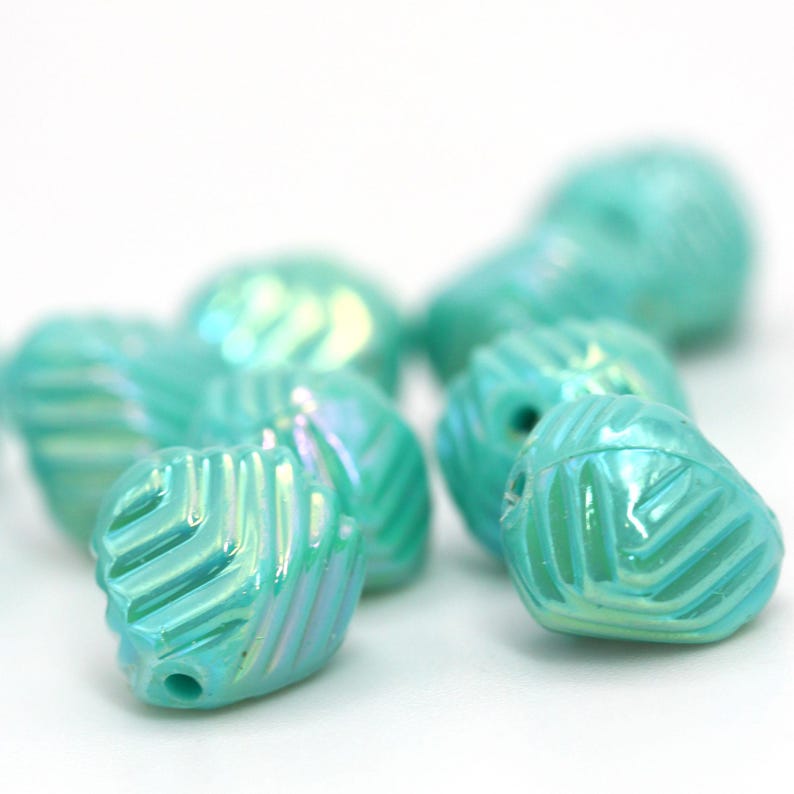 May include: Close-up of several iridescent turquoise leaf-shaped beads. Each bead has a detailed, textured surface with a hole for stringing. The beads are scattered, with some in focus and others blurred, against a white background.