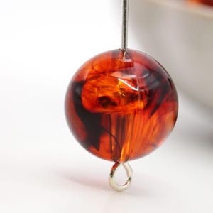 May include: A close-up of a round, translucent orange bead with dark brown swirls. The bead is suspended by a silver-colored wire and has a small silver loop at the bottom. The bead is likely for jewelry making.