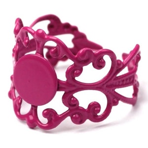 May include: A fuchsia pink metal ring with a filigree design. The ring has a round opening in the center, which is designed to hold a cabochon.