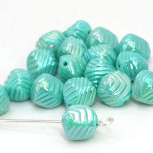 May include: A collection of iridescent turquoise beads with a textured, ridged surface. The beads are irregularly shaped and have a hole for stringing. A silver-colored needle is inserted through one bead.