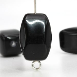 Puede incluir: Primer plano de una cuenta negra brillante con un lazo plateado. La cuenta es rectangular con bordes redondeados. Otras dos cuentas negras están ligeramente desenfocadas en el fondo. Las cuentas son probablemente para la fabricación de joyas.