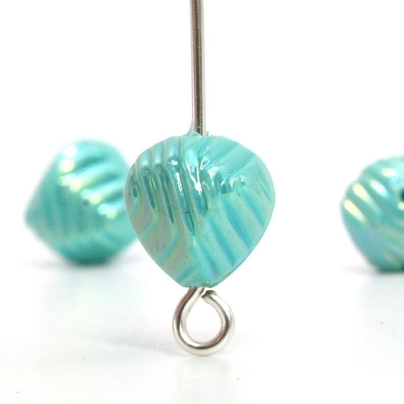 May include: Close-up of three iridescent turquoise glass beads. The beads are heart-shaped with a textured, ridged surface. One bead is attached to a silver wire loop. The background is white.