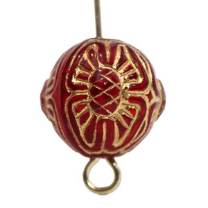 May include: A round, red bead with gold filigree detailing. The bead has a central floral design and is attached to a gold-colored loop and a pin. The bead's surface is glossy, and the gold accents create a striking contrast.