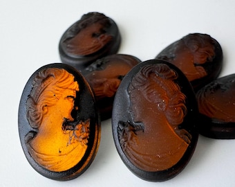 May include: A collection of oval-shaped cameos featuring a relief profile of a person with an intricate hairstyle. The pieces are crafted from translucent amber-coloured glass with a dark, matte black border surrounding the central figure.
