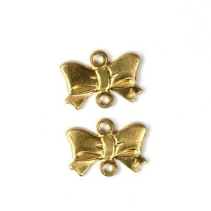 May include: Two gold-colored metal bow charms with two holes each. The bows are detailed and have a three-dimensional look.