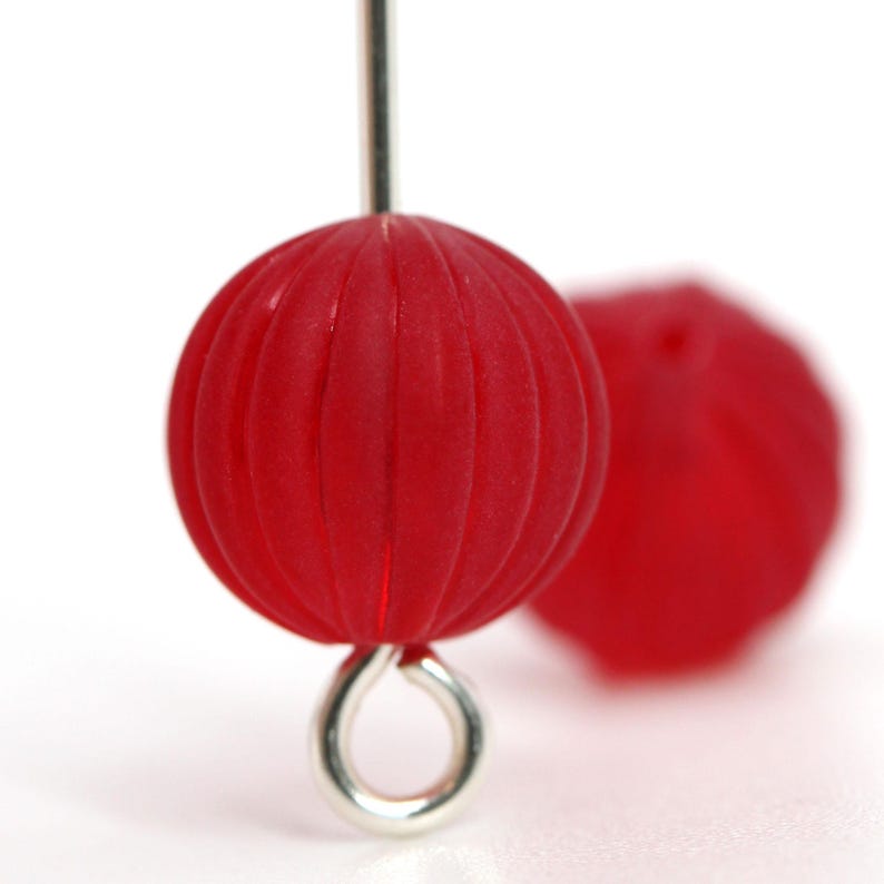 May include: Close-up of a red, round bead with vertical grooves, attached to a silver-colored metal loop and a thin metal rod. Another red bead is blurred in the background. The beads have a matte finish.