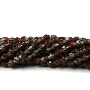 May include: A horizontal strand of faceted beads featuring a mix of deep reddish-brown and translucent grey tones. Each bead has a multi-faceted surface that reflects light, creating a textured appearance across the entire length of the string.