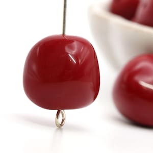 May include: A close-up of a single red cherry-shaped bead with a silver loop attached. The bead is glossy and smooth.