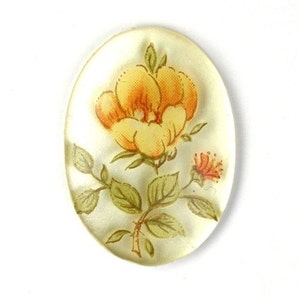 May include: A clear oval-shaped cabochon with a yellow flower and green leaves painted on it.