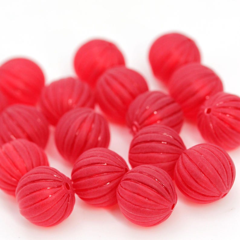 May include: A collection of red, round beads with a grooved surface. The beads are scattered against a white background. Each bead has a small hole, suggesting they are for stringing. The beads are a vibrant red color.
