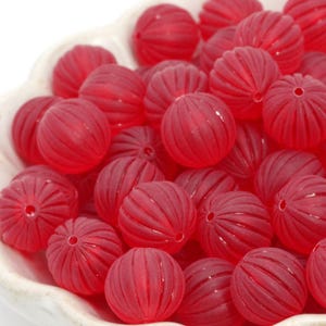 May include: A collection of translucent red beads with a ribbed, spherical design. Each bead has a small hole for stringing. The beads are displayed in a white, scalloped-edge bowl, creating a visually appealing arrangement for crafting projects.
