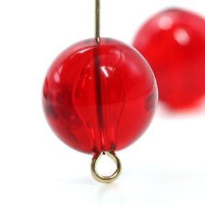 May include: Close-up of two translucent red beads. The front bead is in focus, showing a gold-colored loop at the bottom and a gold wire running through the center. The second bead is slightly out of focus in the background.