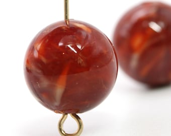 Può includere: Primo piano di due perline rotonde rosse marmorizzate con finitura lucida. Ogni perlina è attaccata a un anello di metallo color oro e a un'asta sottile di metallo color oro. Le perline hanno un motivo a spirale di rosso più chiaro e bianco.