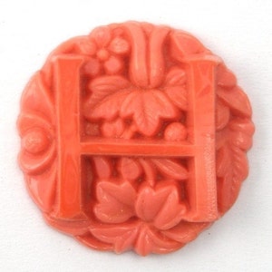 May include: A coral-colored plastic button with a raised letter "H" surrounded by a floral design.