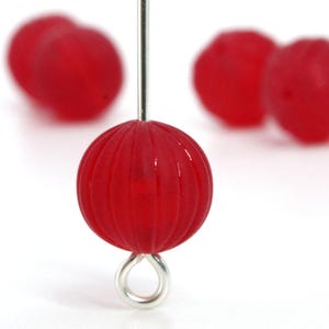 May include: Close-up of a red, round bead with vertical grooves, suspended by a silver-colored metal pin and loop. Several other blurred red beads are in the background, all against a white backdrop. The bead has a glossy finish.