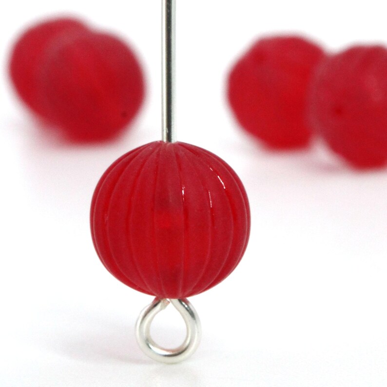 May include: Close-up of a red, round bead with vertical grooves, suspended by a silver-colored metal pin and loop. Several other blurred red beads are in the background, all against a white backdrop. The bead has a glossy finish.