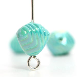 May include: Close-up of a turquoise glass bead with a spiral design and a silver-colored eye pin. The bead is a light blue-green color with a glossy finish. Another bead is slightly out of focus in the background.