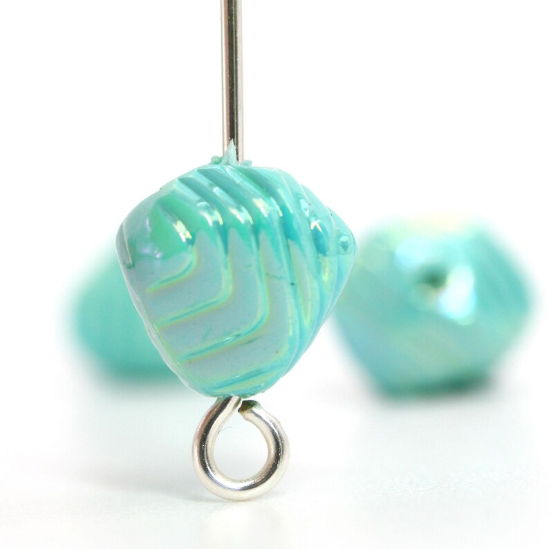 May include: Close-up of a turquoise glass bead with a spiral design and a silver-colored eye pin. The bead is a light blue-green color with a glossy finish. Another bead is slightly out of focus in the background.