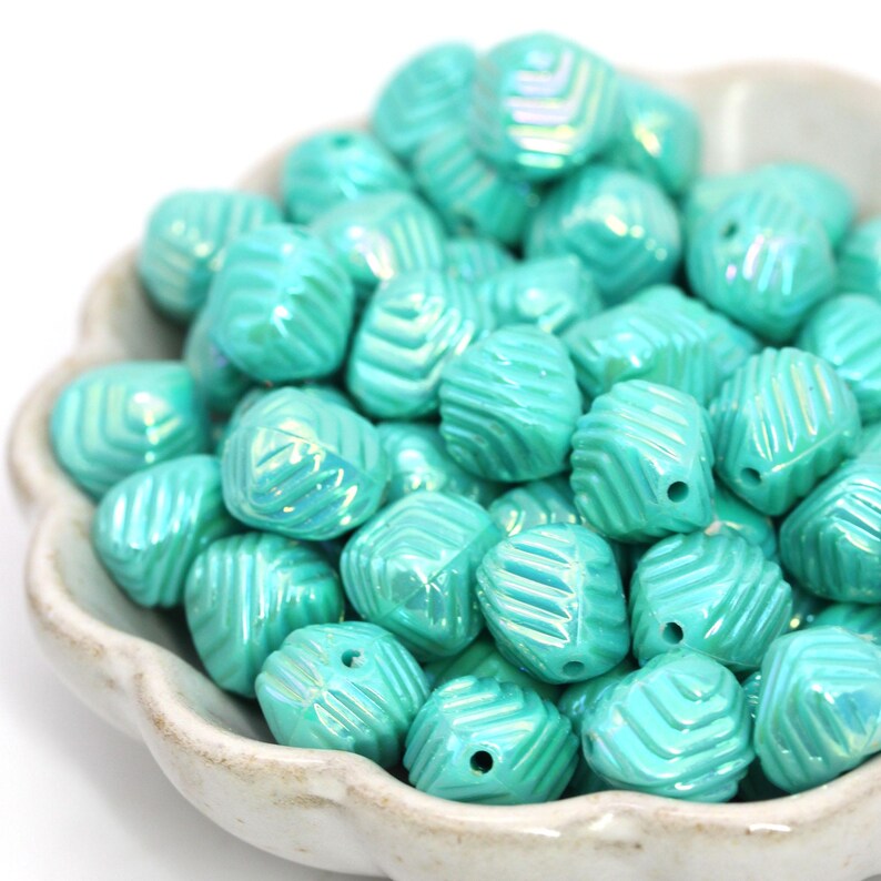 May include: A collection of turquoise-colored beads with a textured, ridged design. The beads are in a white, scalloped-edge bowl. The beads have a glossy finish and are suitable for jewelry making or craft projects.