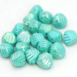 May include: A collection of iridescent turquoise beads. Each bead has a geometric pattern of diagonal lines and a hole for stringing. The beads are scattered on a white surface, creating a visually appealing arrangement.