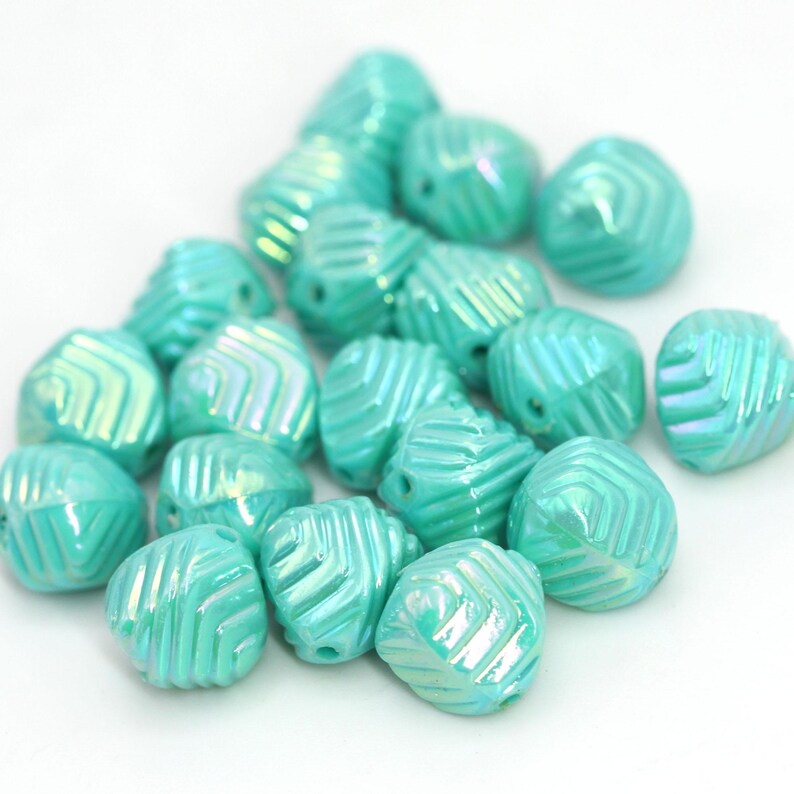 May include: A collection of iridescent turquoise beads. Each bead has a geometric pattern of diagonal lines and a hole for stringing. The beads are scattered on a white surface, creating a visually appealing arrangement.