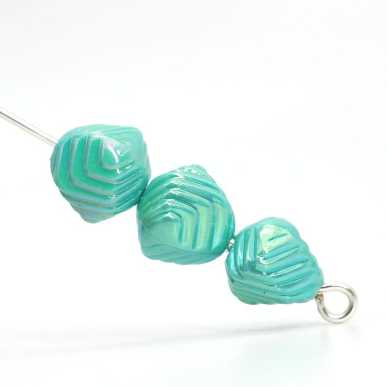 May include: Three iridescent turquoise glass beads with a geometric pattern. The beads are strung on a silver-colored wire and a metal loop. The beads have a textured surface and a pearlescent sheen.