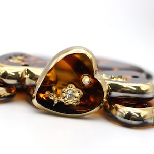 May include: Close-up of heart-shaped hair clips with a tortoiseshell pattern and gold trim. Each clip features a small gold flower with a central clear rhinestone. The clips are arranged on a white surface.