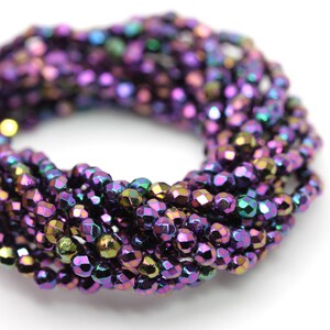 May include: Close-up of a strand of faceted beads in shades of purple, green, and gold. The beads have a metallic sheen and are arranged in a circular pattern against a white background. The beads are small and uniform in size.