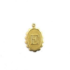 May include: Gold-colored pendant with the letter "E" inside an oval frame. The frame has a textured background and a scalloped edge. A small loop is at the top for attaching to a chain or bracelet. The pendant is on a white background.