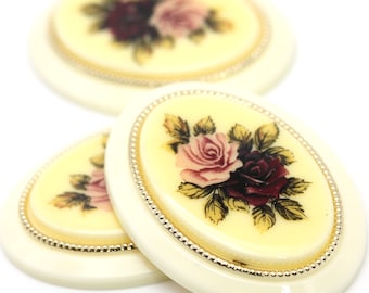 May include: Ivory-colored oval cabochons with a floral rose design. The roses are in shades of pink and burgundy, with green leaves. Each cabochon has a gold-colored beaded border. The cabochons are ideal for jewelry making or craft projects.