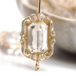 May include: A clear, rectangular glass bead with a gold-colored ornate frame. The bead is suspended on a gold-colored wire, showcasing its intricate design and vintage aesthetic. The bead's shape is reminiscent of a small, decorative window.
